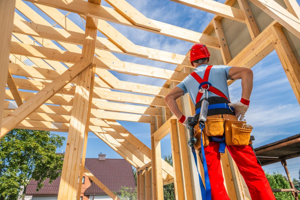 A worker in a red hard hat and safety harness standing inside a timber-framed house, looking up at exposed roof trusses against a blue sky.