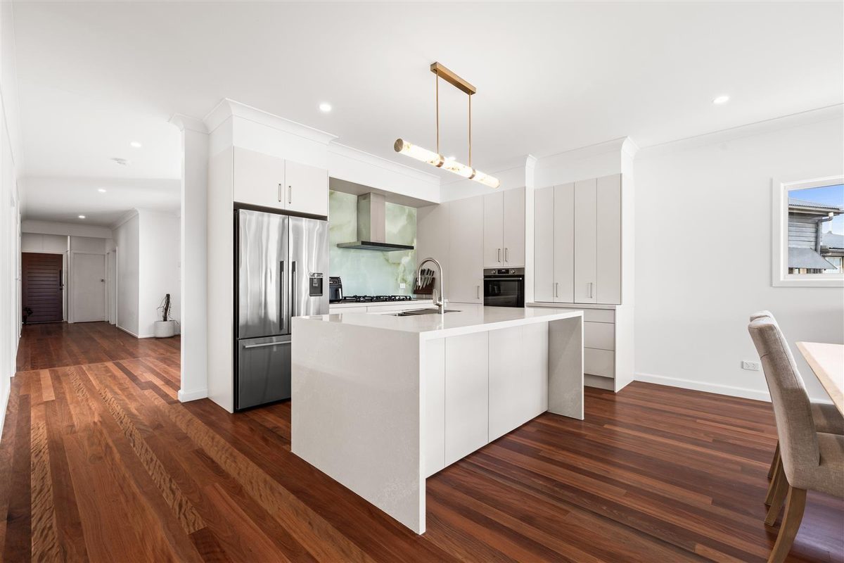 A bright modern kitchen with a white waterfall island, stainless steel refrigerator and polished timber floors.